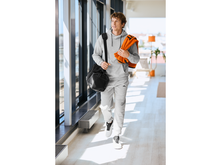 Mees hallis kapuutsiga dressipluusis ja dressipükstes, musta spordikoti ja oranži rätikuga.