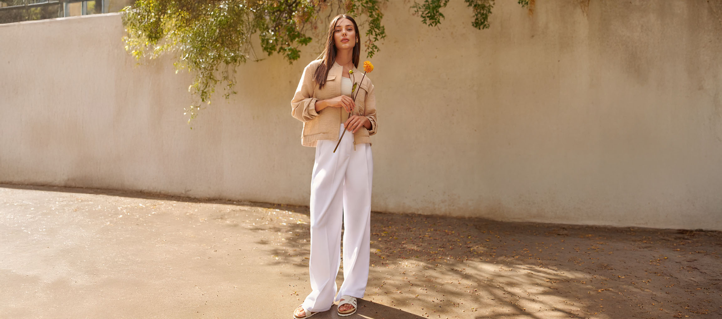 Женщина в бежевой куртке, белых брюках и сандалиях, держит желтый цветок.