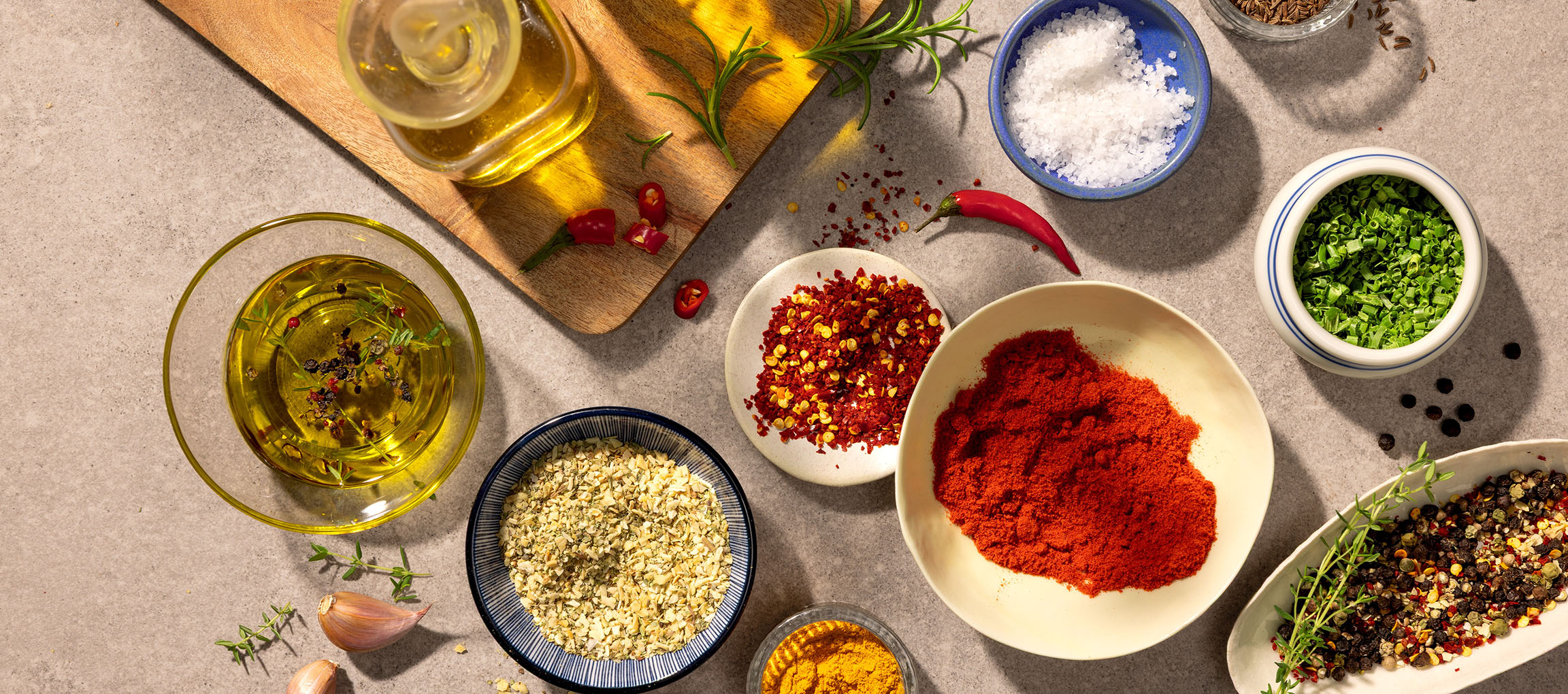 Erinevad vürtsid ja õlid, sealhulgas paprika, tšilli, sool, pipar ja oliiviõli, hallil taustal.