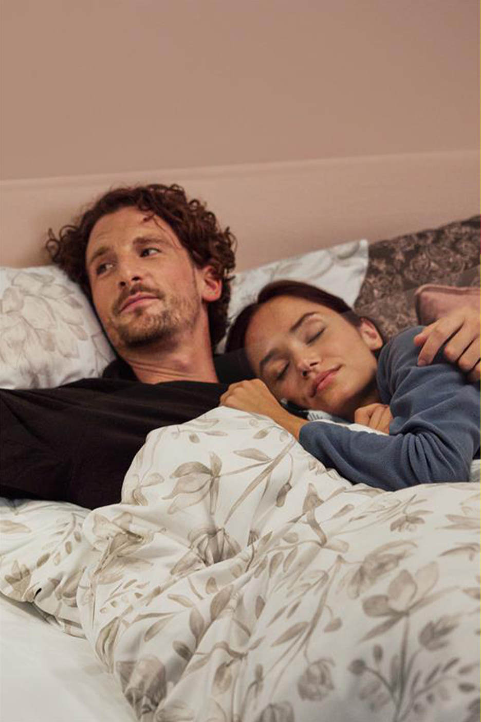 Paar voodis, mees on ärkvel, naine magab, neil on peal lillemustriga tekk.