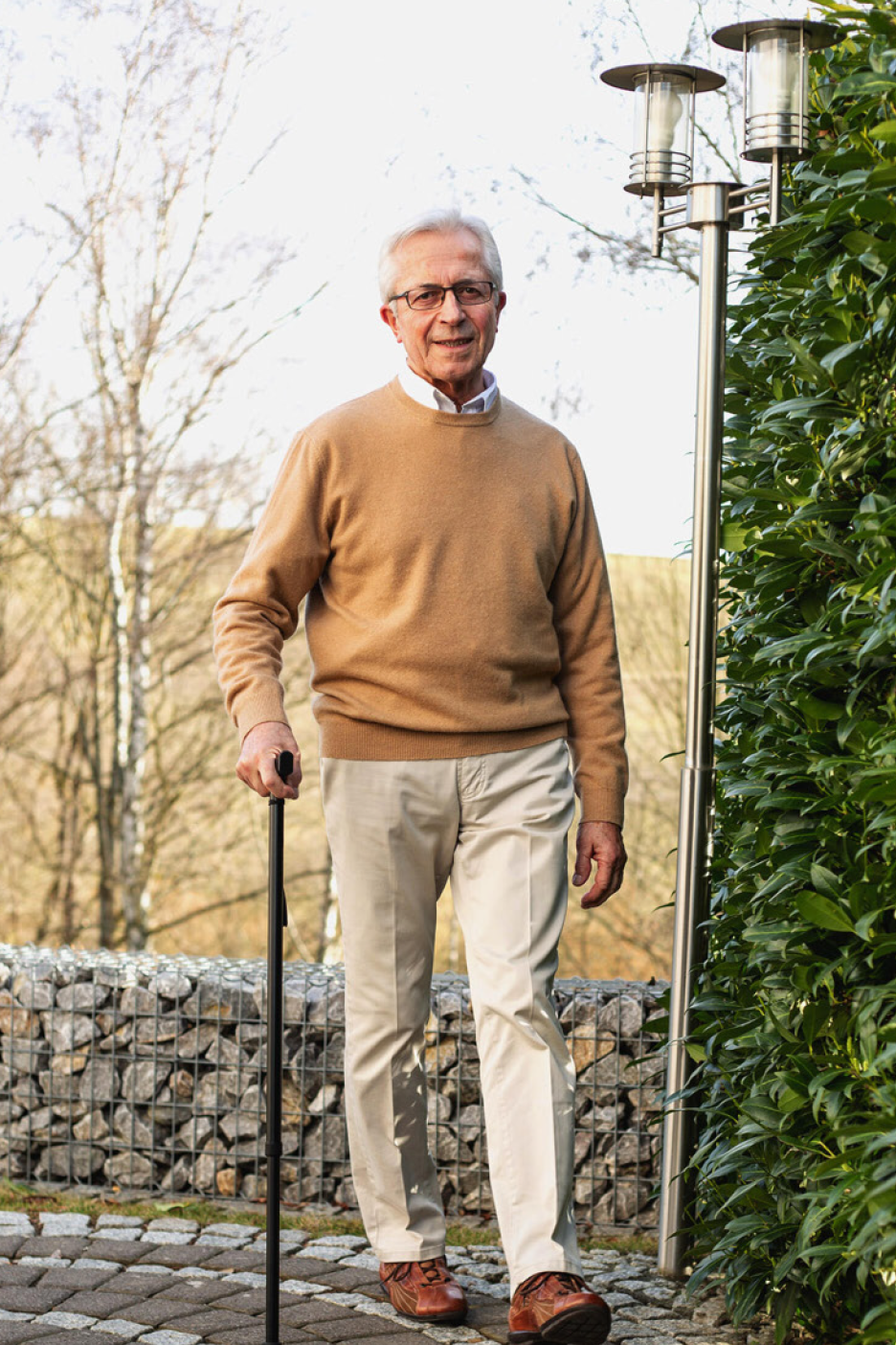 Eakas mees beežis kampsunis ja heledates pükstes, toetudes kepile, õues.