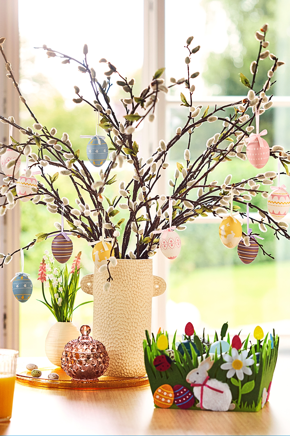 Pajuokstest ja lihavõttemunadest kaunistus laual, taustal aken ja roheline muru.