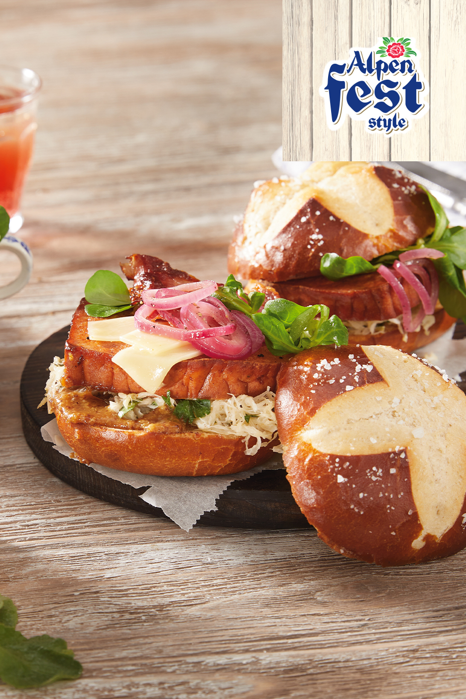 Kaks soolase kringli kuklit lihaleiva, juustu, hapukapsa ja marineeritud sibulaga, taustal jook.