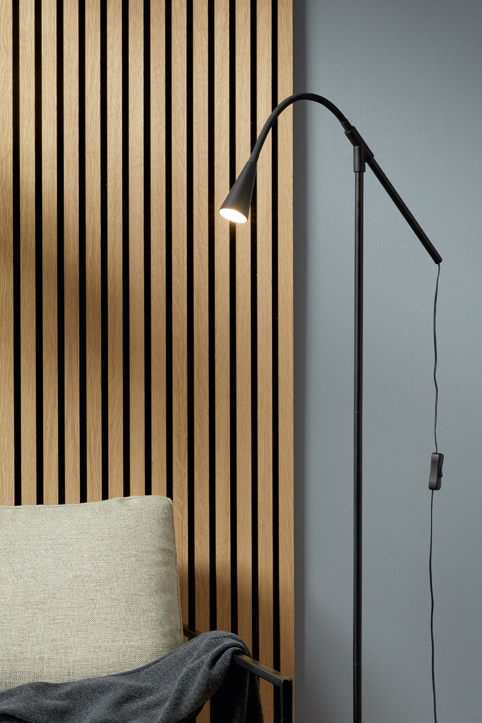 Must põrandalamp kumera varre ja valgustatud lambiga puitpaneeli taustal.