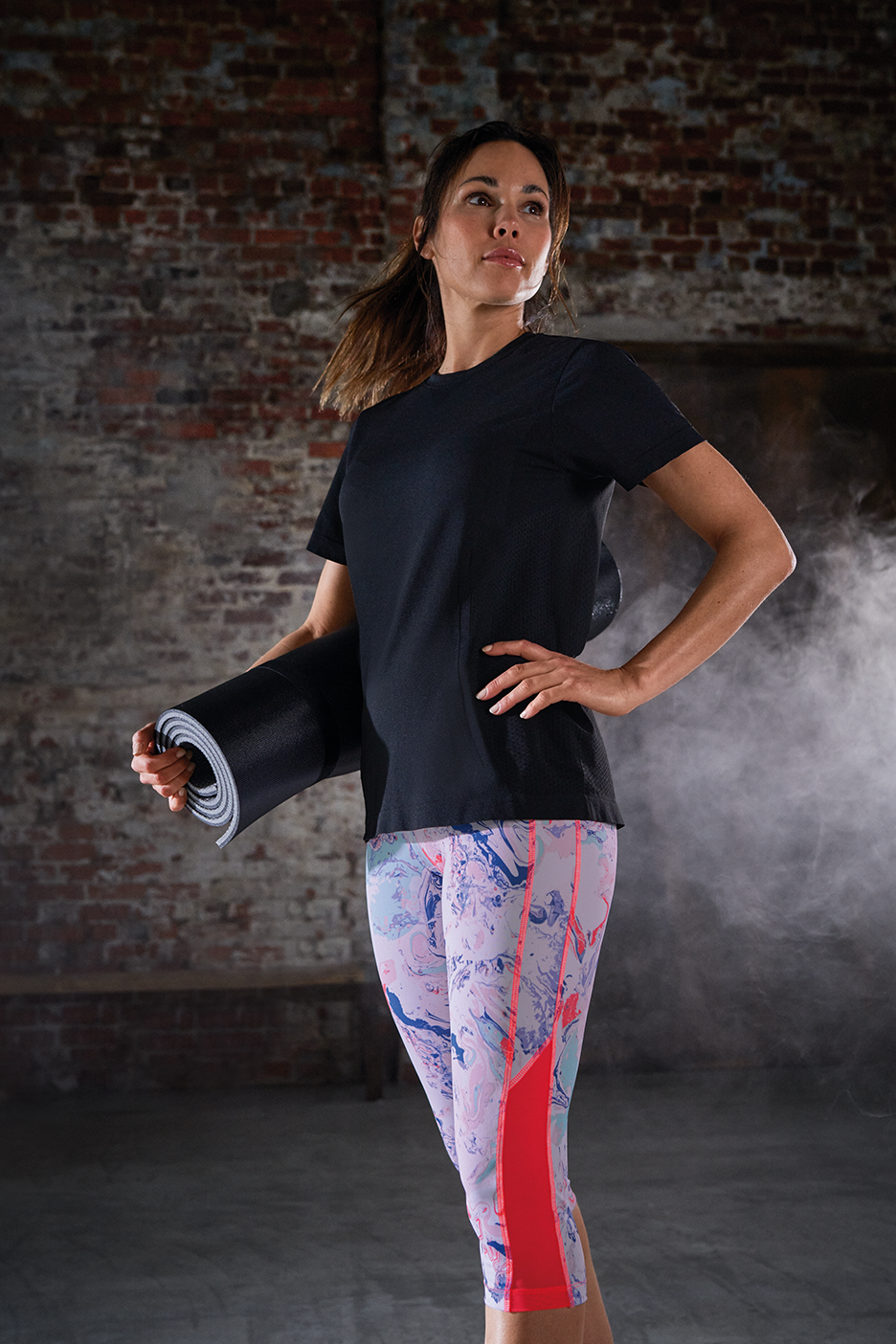 Naine mustas spordisärgis ja värvilistes retuusides, joogamatt käes, treeningruumis
