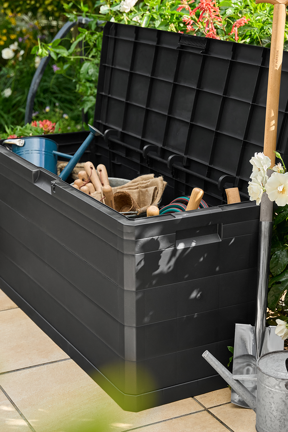 Must aiahoiukast aiatööriistade, kastekannu ja labidaga terrassil, taustal rohelised taimed.