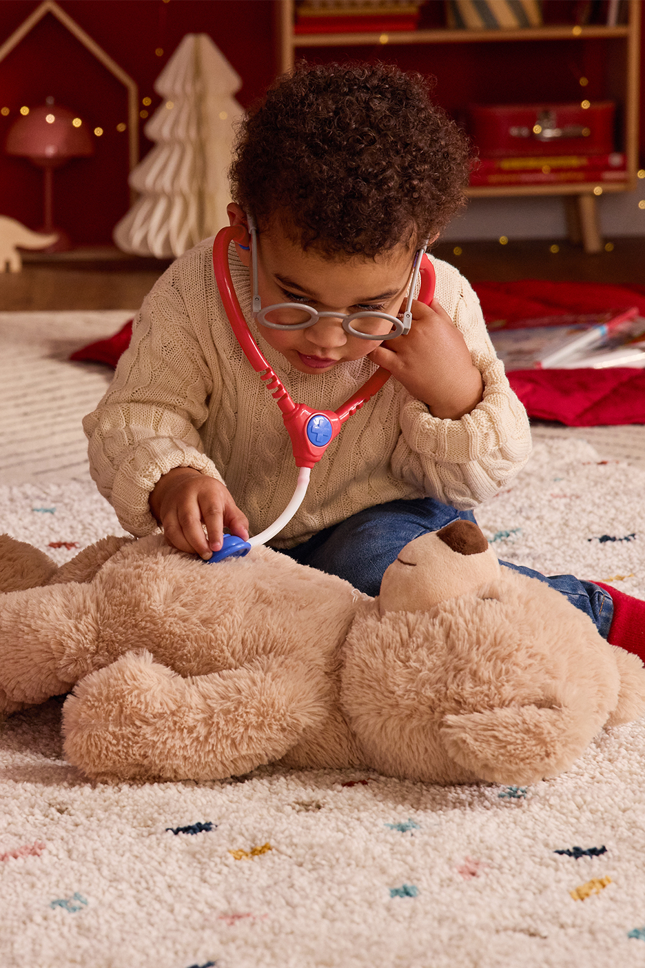 Väikelaps kampsunis ja teksades mängib kaisukaruga arsti, istudes vaibal.