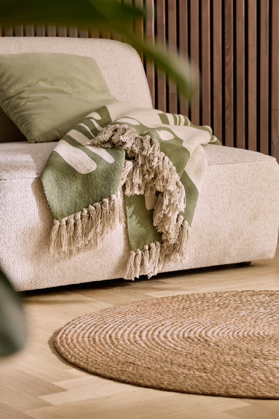 Rohelise mustriga pleed ja padi beežil diivanil, heledal puitpõrandal on ümmargune vaip.