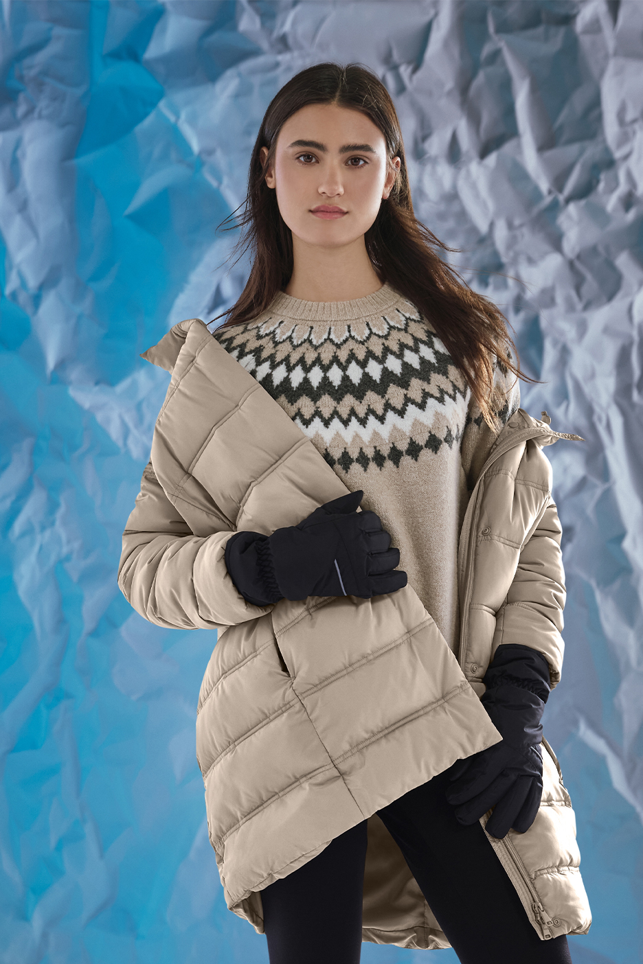 Naine beežis tepitud jopes, mustrilises kampsunis ja mustades kinnastes sinisel taustal.