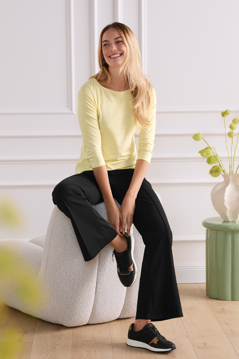 Naine kollase pika varrukaga pluusi ja mustade pükstega istumas heledal diivanil.