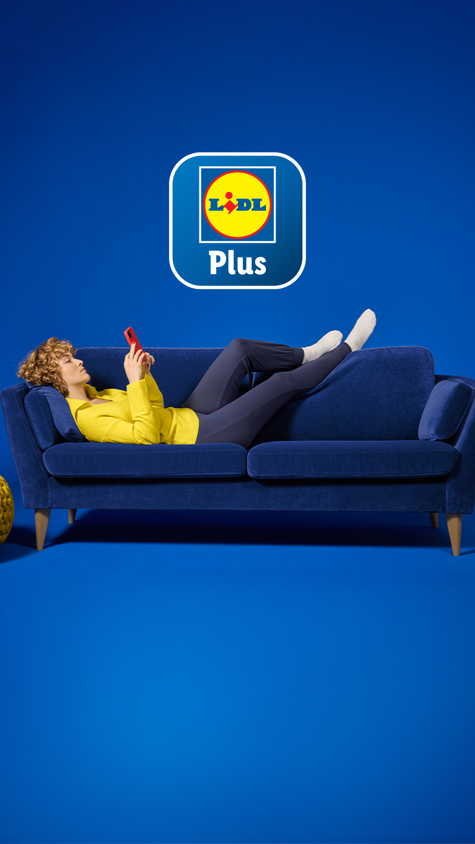 Naine lamab sinisel diivanil, kasutades telefoni, Lidl Plus logo kohal.