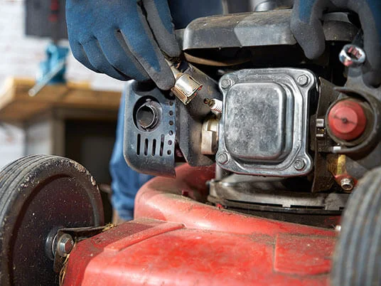 Руки в перчатках заменяют свечу зажигания на красной газонокосилке.