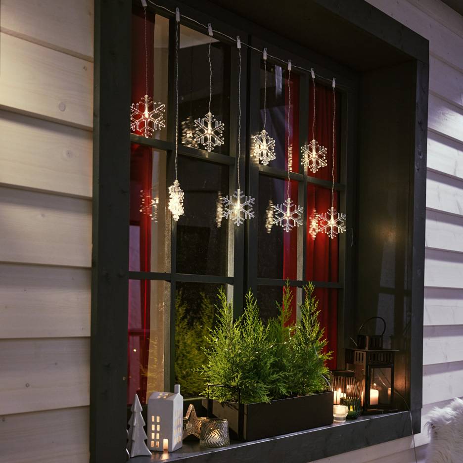 Lumehelbekujulised valgusketid aknas väikeste kuuskede ja küünaldega aknalaual.