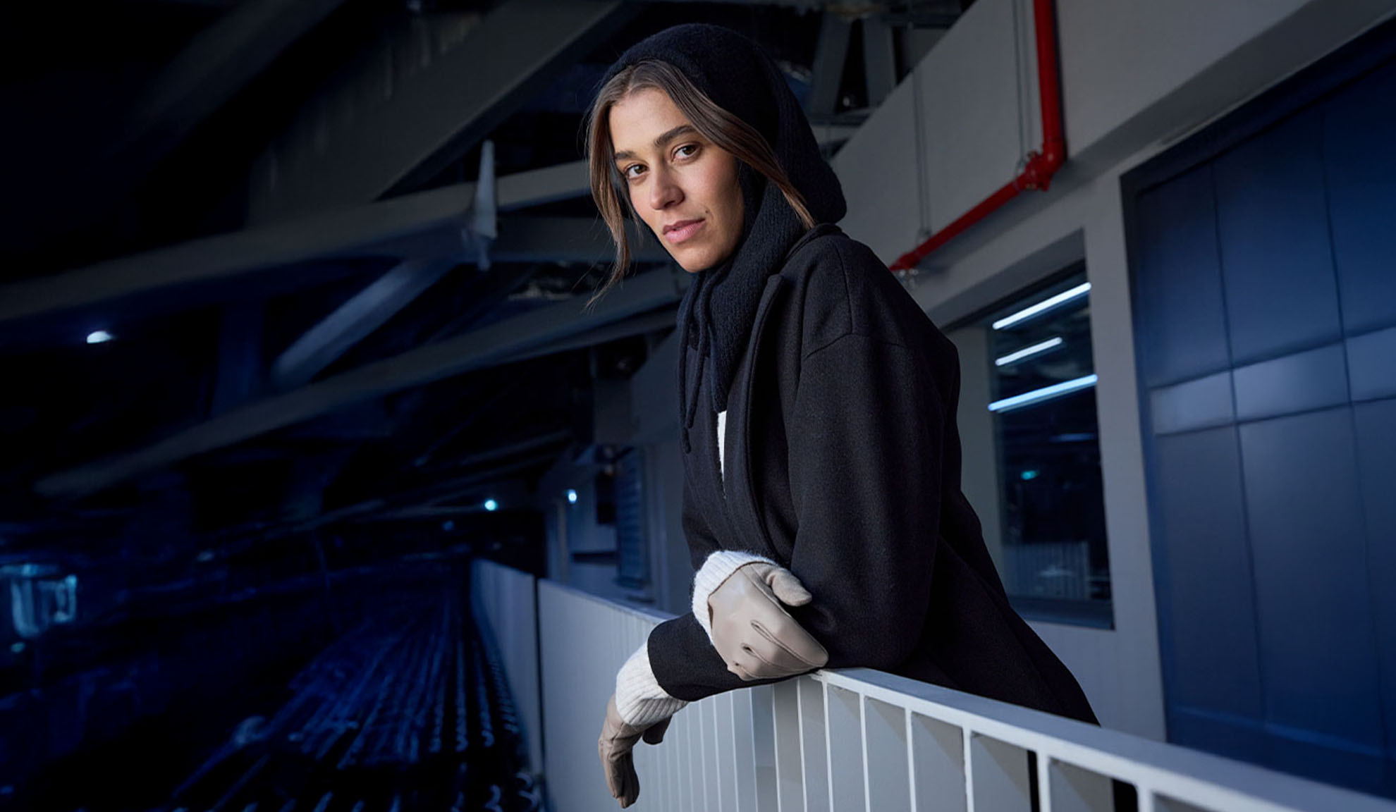 Naine mustas mantlis ja kapuutsiga salliga, toetudes piirdele kaasaegses keskkonnas.