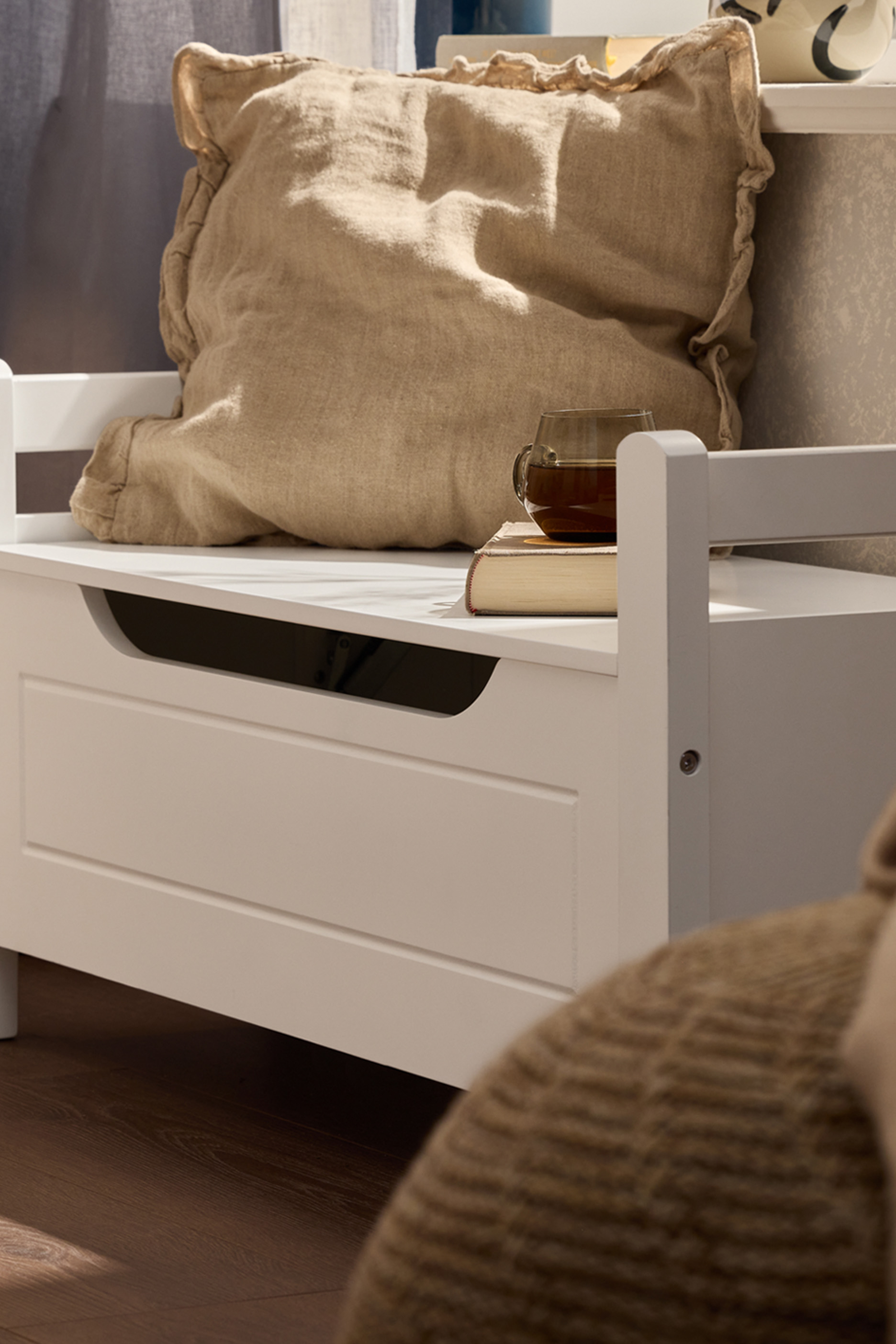 Белая скамья для хранения с подушкой, книгой и чашкой чая в уютной комнате.