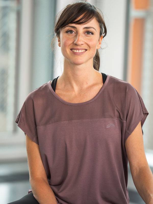 Улыбающаяся женщина в фиолетовом спортивном топе с короткими рукавами.