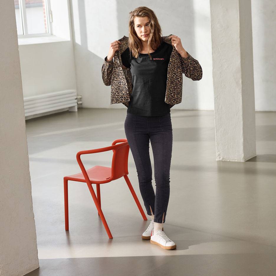 Женщина в черной футболке с надписью, джинсах и куртке с леопардовым принтом, стоящая рядом с красным стулом.