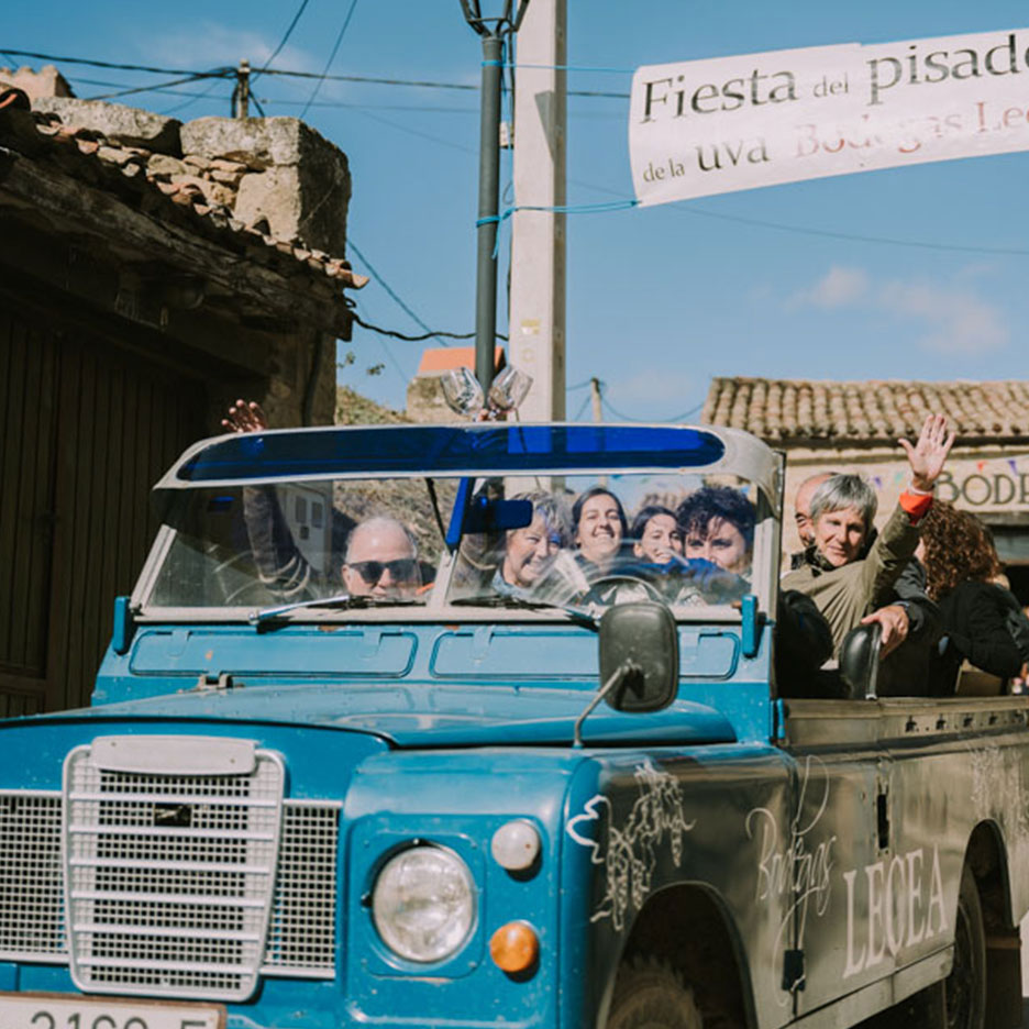 Inimesed lehvitavad sinisest maasturist veinifestivali ajal, bänneril on kiri 'Fiesta del pisado de la uva'.