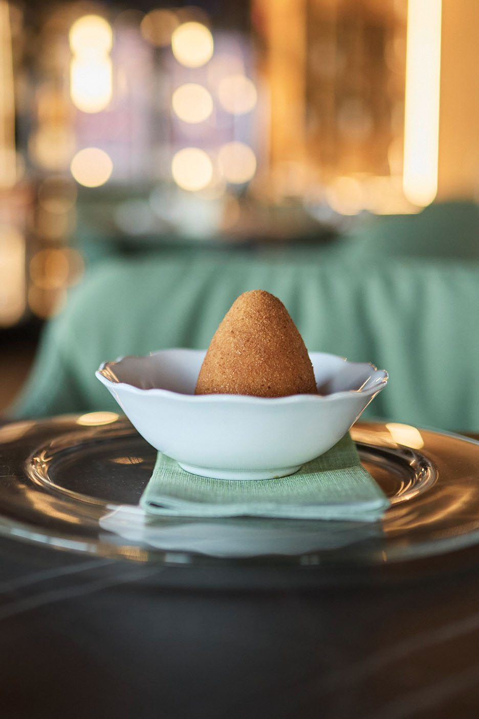 Kuldne arancini valges kausis rohelisel salvrätikul ja klaasplaadil.