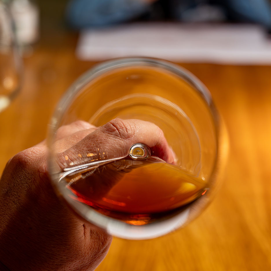 Käsi hoiab klaasi punast veini või merevaigukarva kanget alkoholi puidust laual.