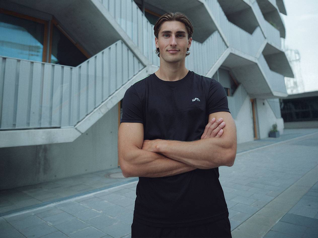 Mees mustas logoga T-särgis ja mustades lühikestes pükstes, seisab õues käed risti.