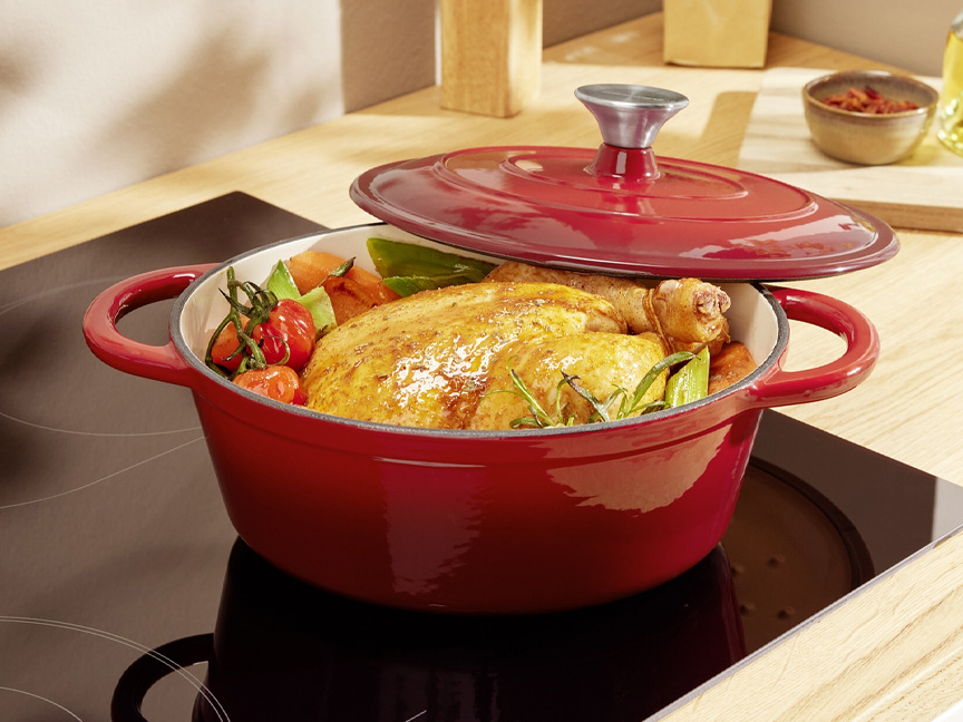 Красная чугунная кастрюля с жареной курицей и овощами на индукционной плите.