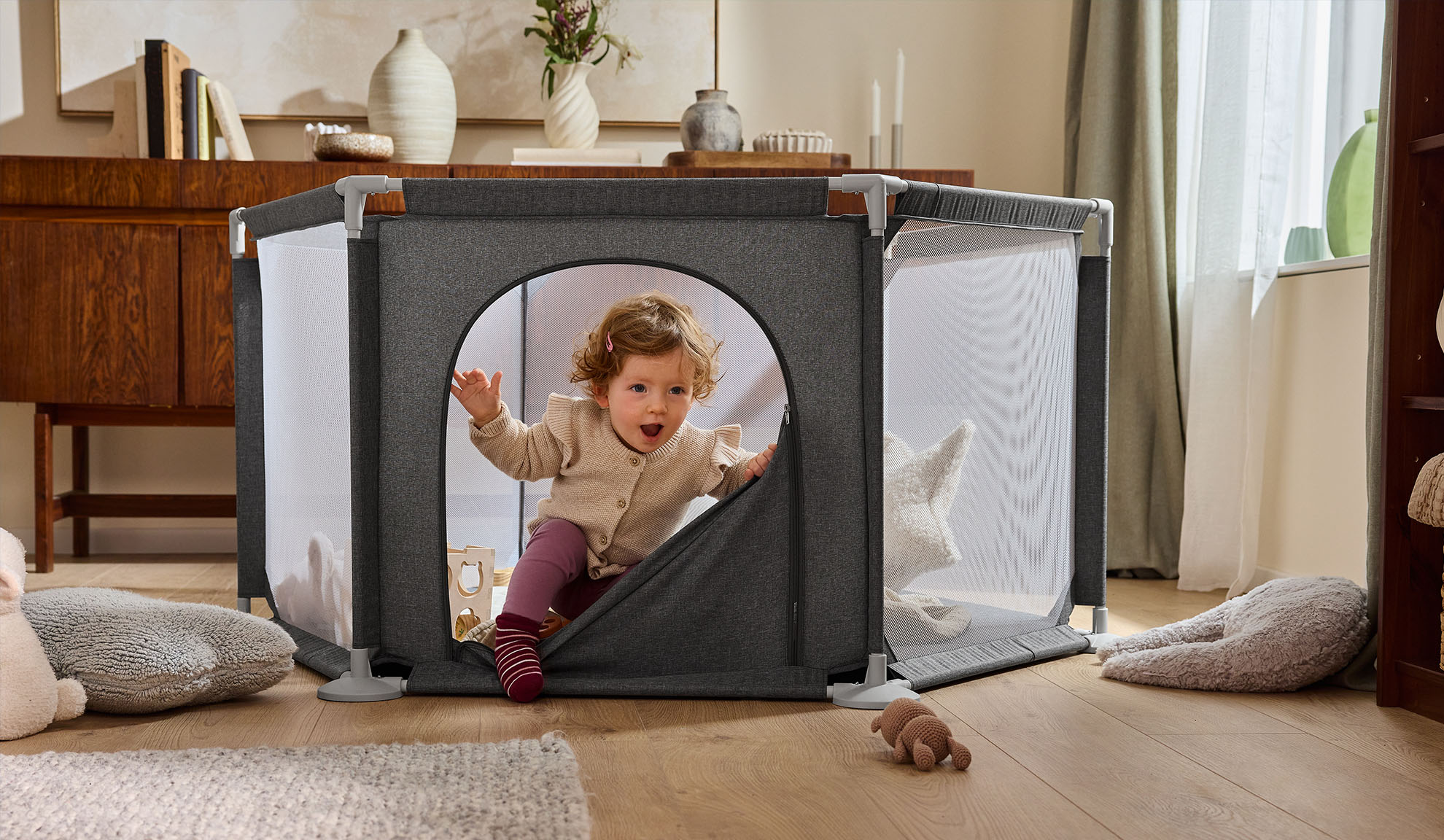 Счастливый малыш вылезает из серого манежа в уютной гостиной.