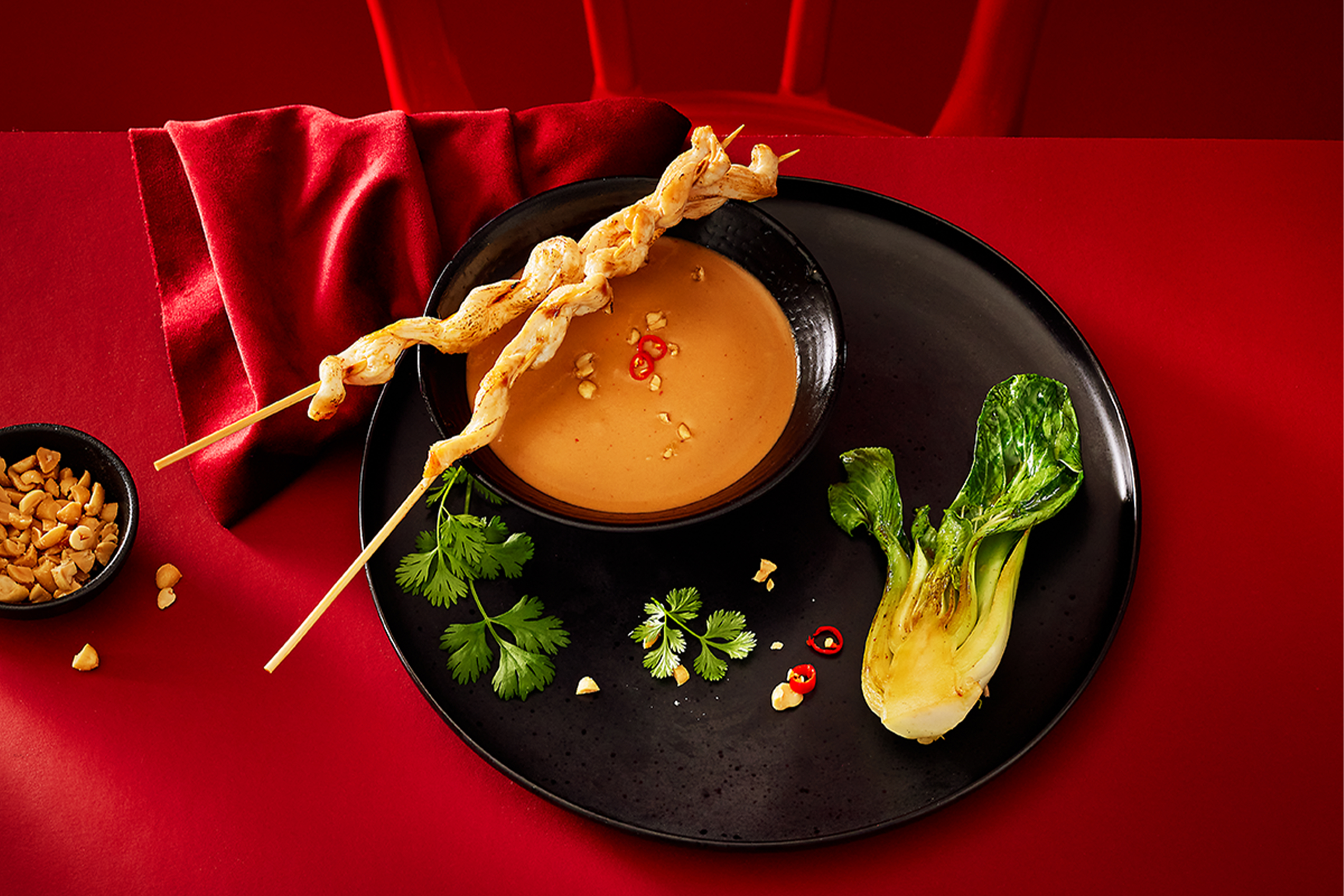 Kana satay varras maapähklikastme, pak choi ja koriandriga punasel taustal.
