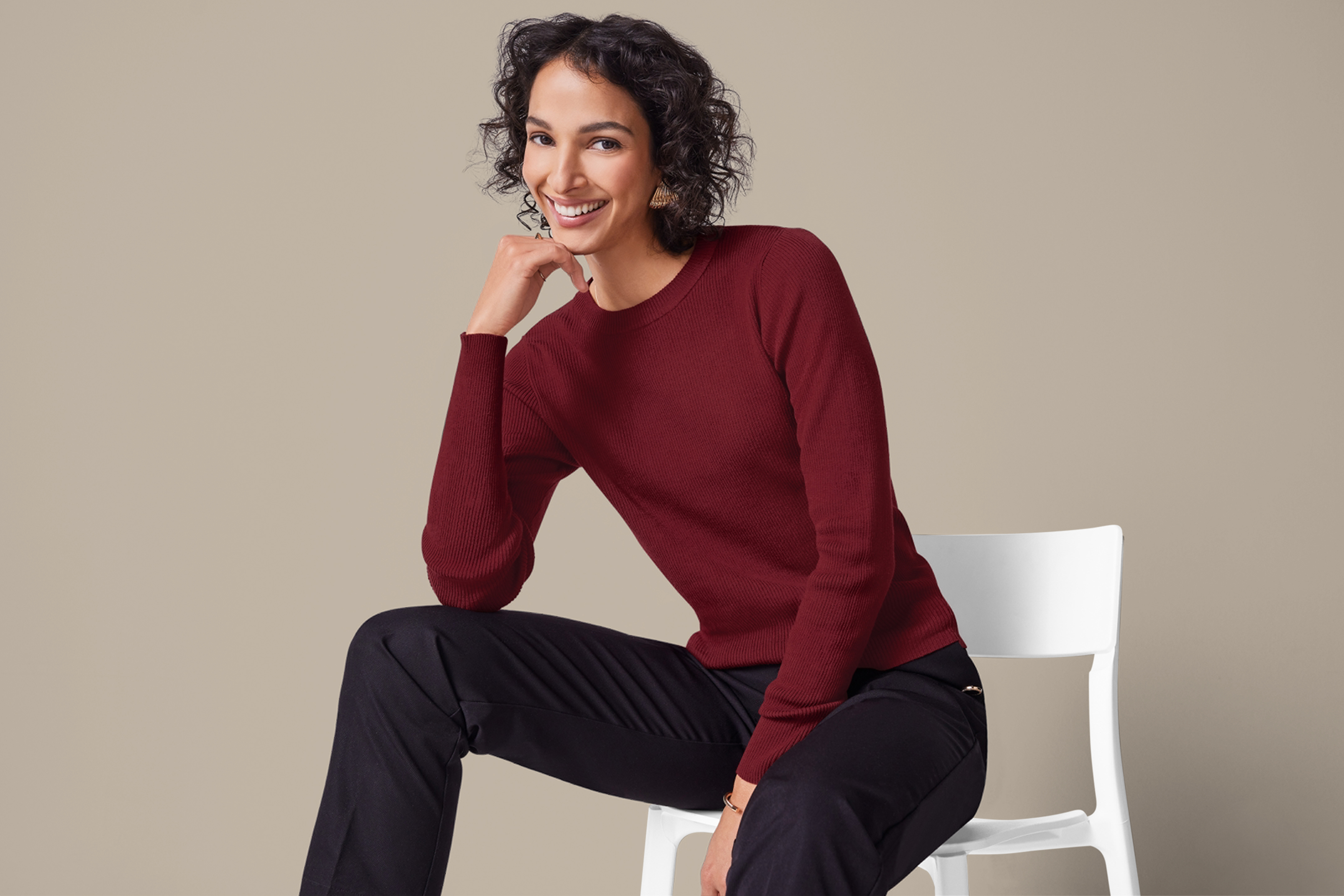 Женщина в бордовом свитере и черных брюках сидит на белом стуле.