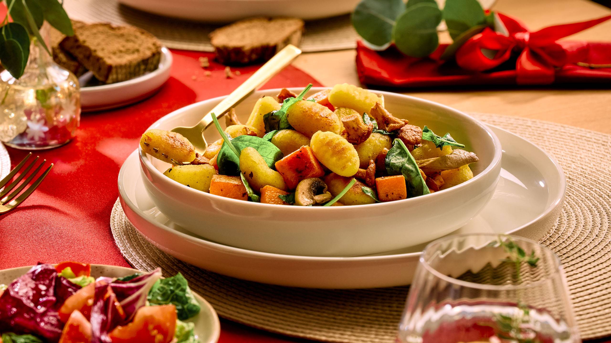 Kauss gnocchi, köögiviljade ja seentega, salat ja leib punasel laual.