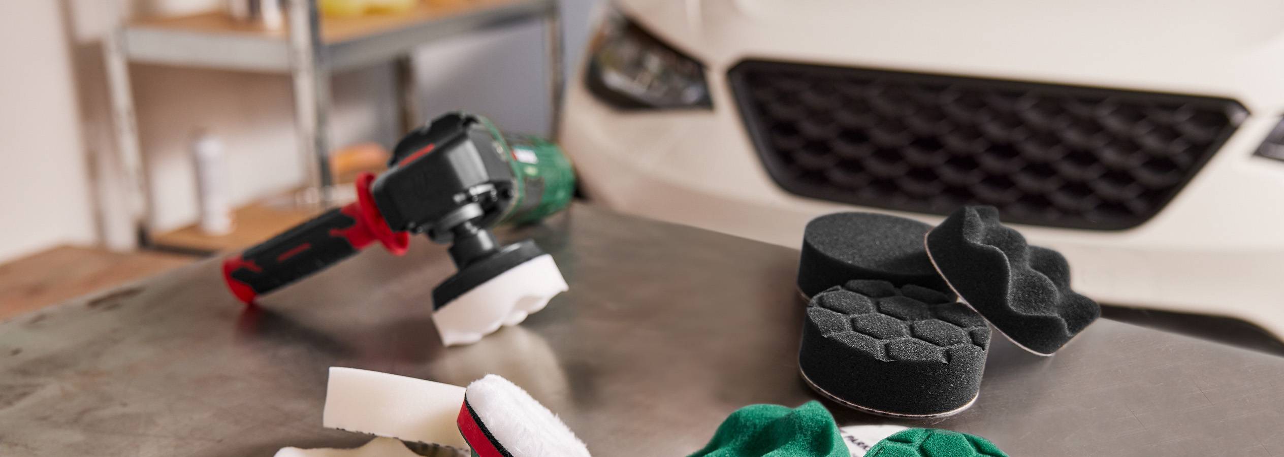 Poleerimismasin ja erinevad poleerimiskäsnad laual valge auto ees.
