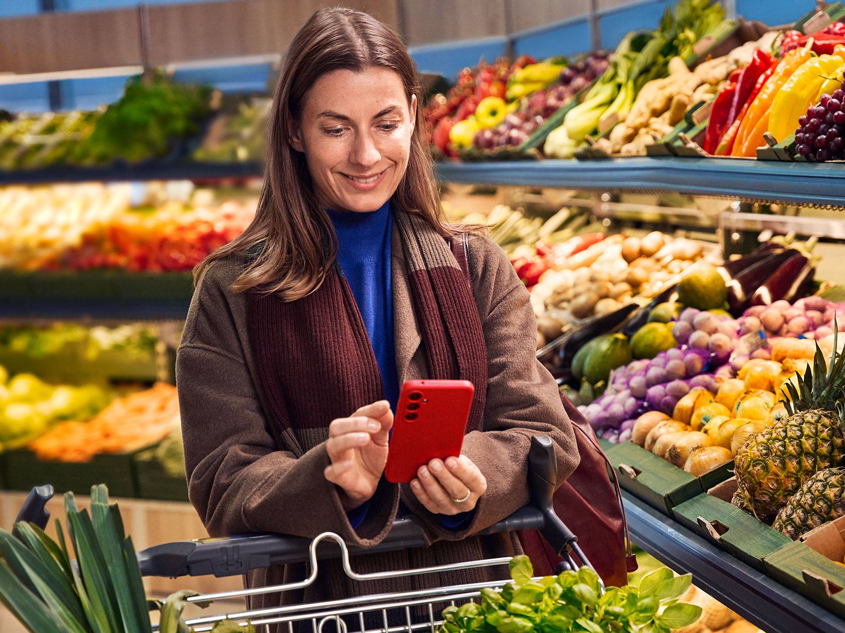 Naine vaatab supermarketis telefonist, ostukäru täis köögivilju ja puuvilju.
