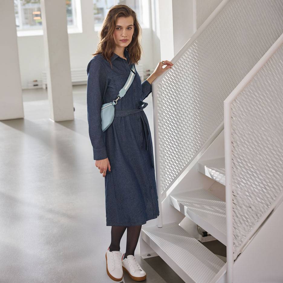 Женщина в джинсовом платье-рубашке, черных колготках и белых кроссовках, со светло-голубой поясной сумкой.